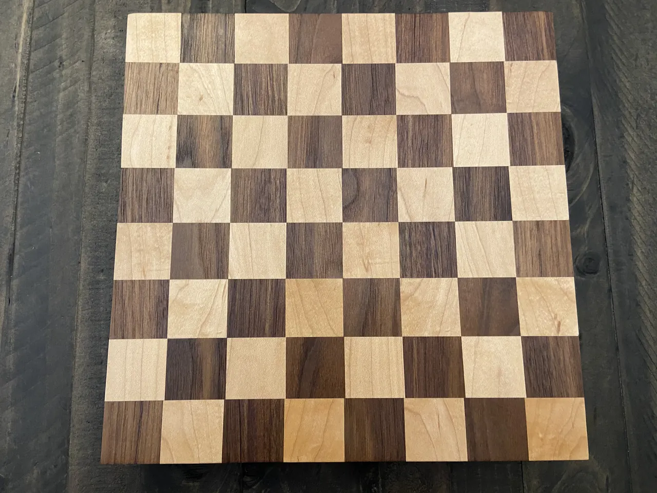 A finished end-grain cutting board in a maple and walnut checkerboard pattern, resting on dark planks.